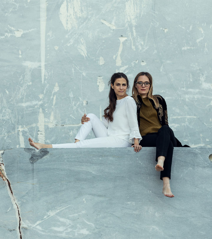 Portrait of two women seated barefoot on a large grey marble block in a quarry, blending natural stone textures with modern design sensibility—symbolizing OnEntropy’s creative partnership.