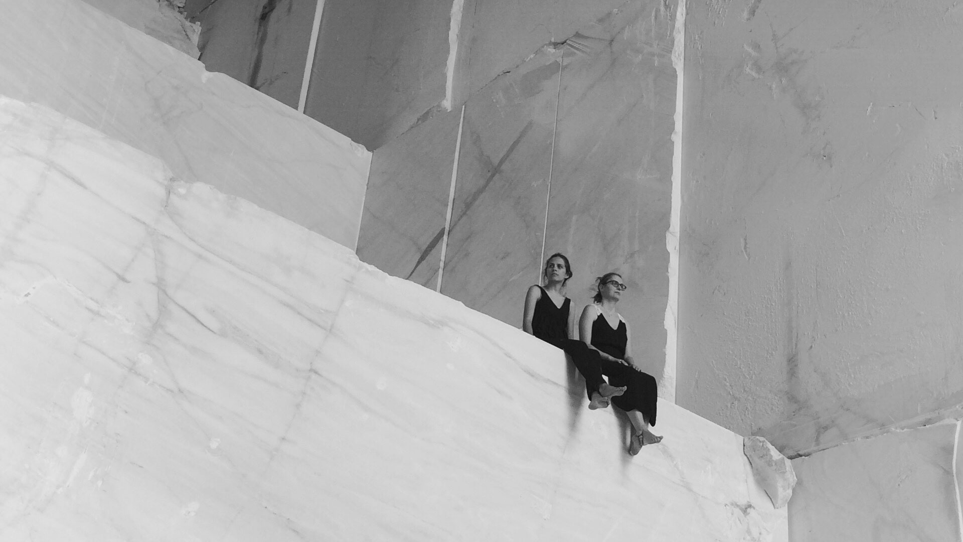 Two women in black sitting on a large marble block inside a quarry, captured in a minimal black and white composition highlighting scale, materiality, and human presence in stone architecture.
